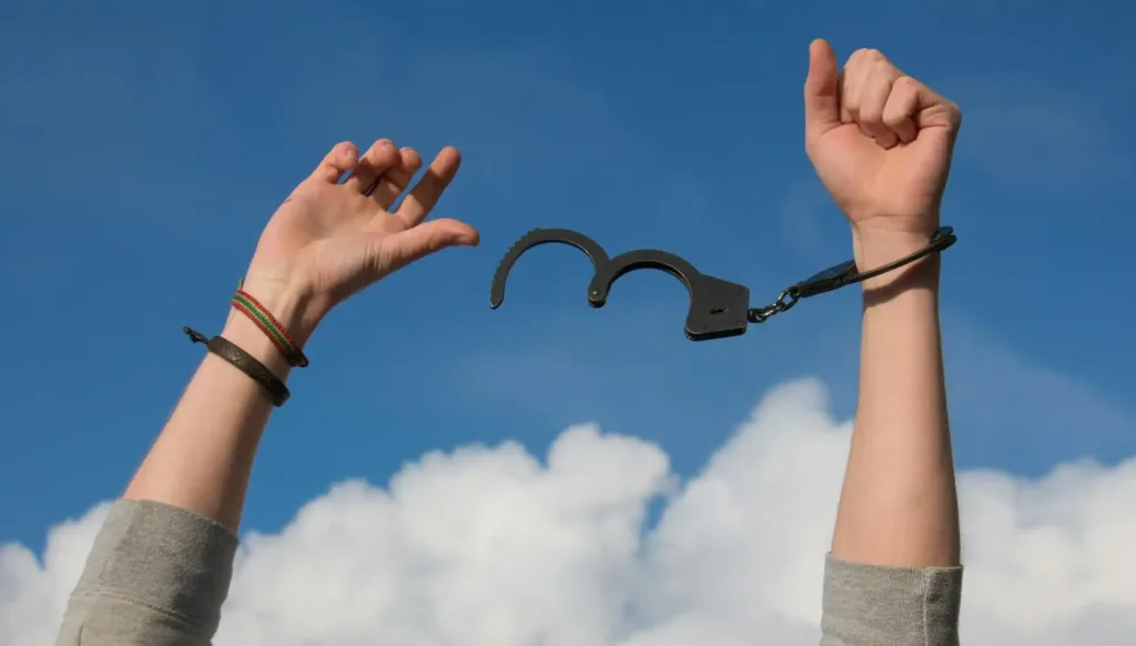 Dos brazos en alto, el de la derecha con unas esposas puestas y con la mano cerrada y el de la izquierda con la mano medio abierta y sin esposa.